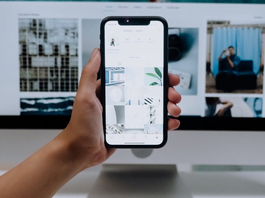Persona sosteniendo un móvil con un perfil de Instagram minimalista, mientras se ve de fondo una pantalla con imágenes digitales.